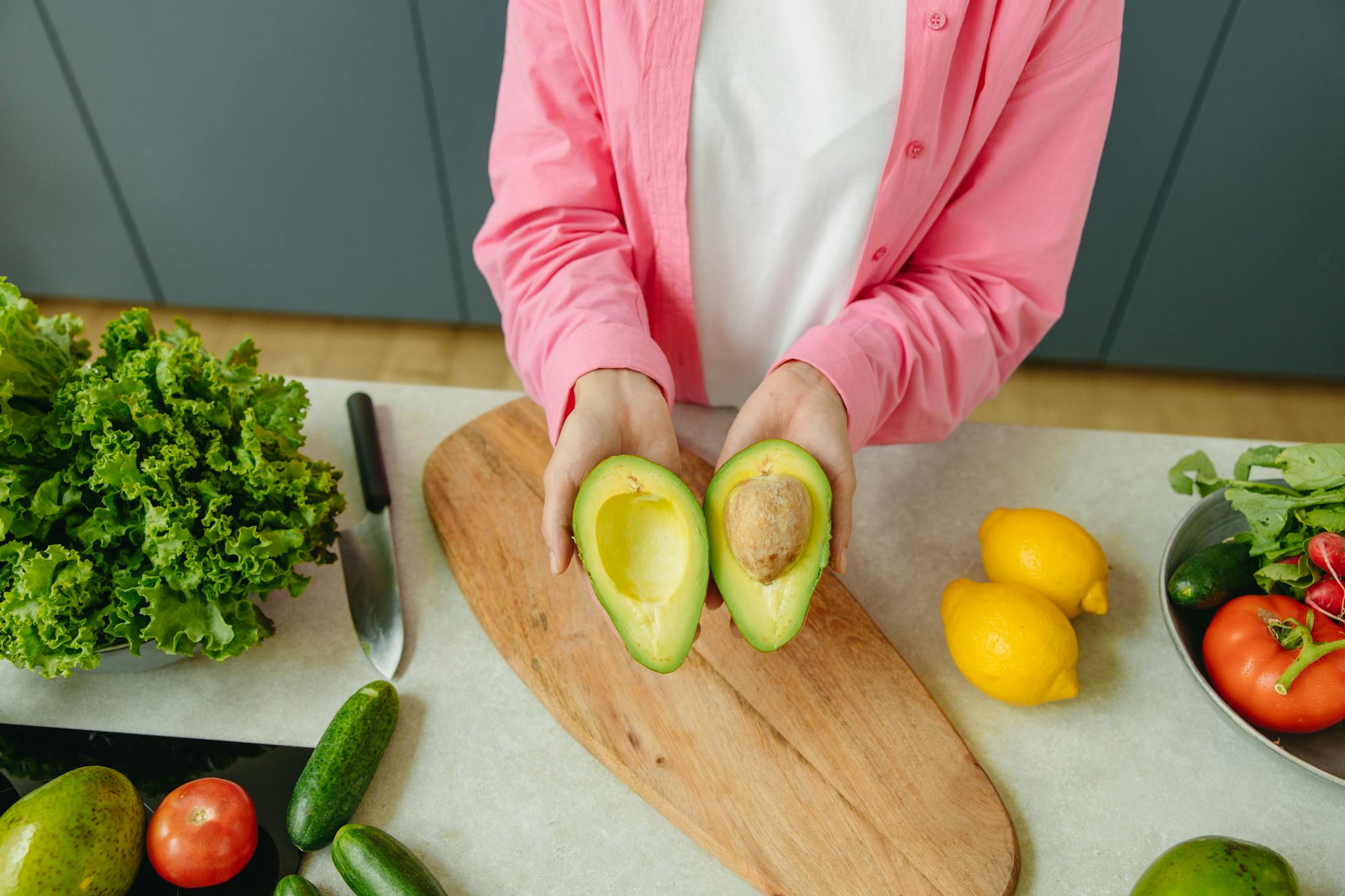 Person holding a sliced avocado on a kitchen counter with fresh vegetables around.