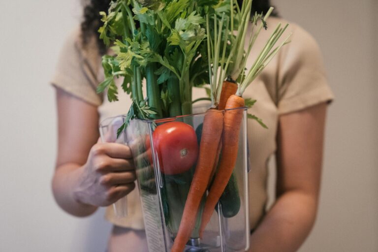 Woman holding a blender pitcher filled with fresh vegetables like carrots, tomato, and celery, promoting healthy eating.